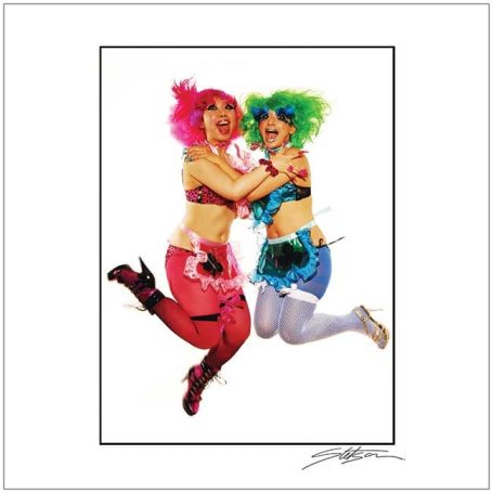 Two vibrant performers with colourful hair and outfits jumping joyfully, smiling at the camera.
