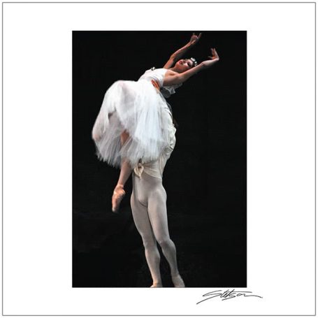 A ballet dancer lifts another dancer wearing a white tutu against a dark background.