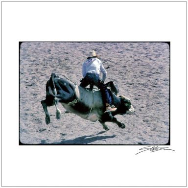 A cowboy riding a bucking bull in a sandy arena.