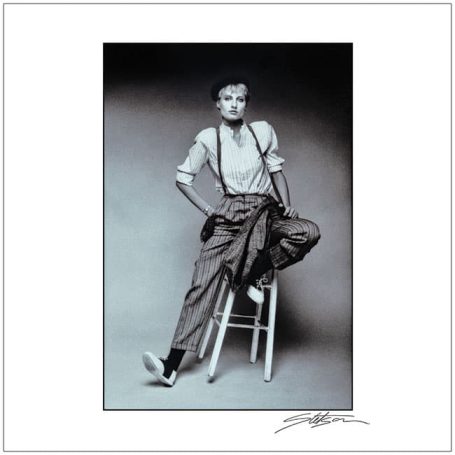 A person sitting on a stool, wearing suspenders and baggy trousers in black and white.