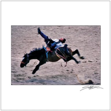 A rider falls from a horse during a rodeo event on sandy ground.