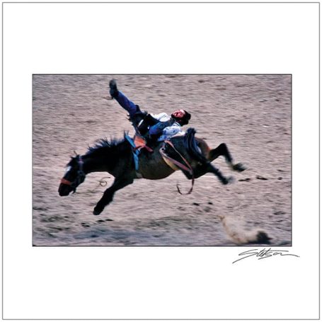 A rider falls from a horse during a rodeo event on sandy ground.