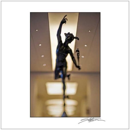 Bronze sculpture of a figure with one arm raised, set against a blurred interior background.