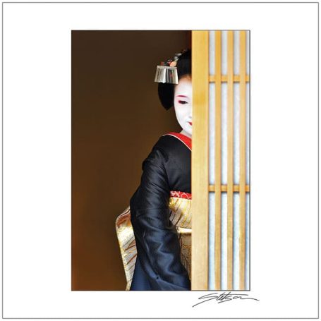 A geisha partially hidden behind a wooden screen, wearing a black and gold kimono.