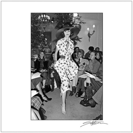 Model in a polka dot dress walking confidently on a runway, with an audience watching.