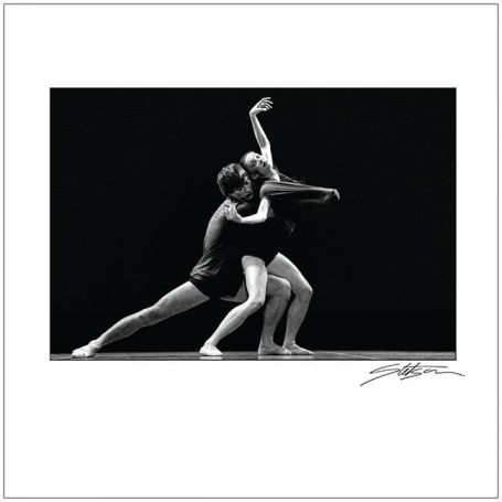 Two dancers perform a dynamic embrace on a dark stage, captured in black and white.