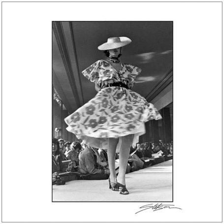 Model walking on runway in a floral dress and wide-brimmed hat during a fashion show.