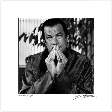 A man in a dark robe sits with hands clasped, looking thoughtfully with a neutral expression.