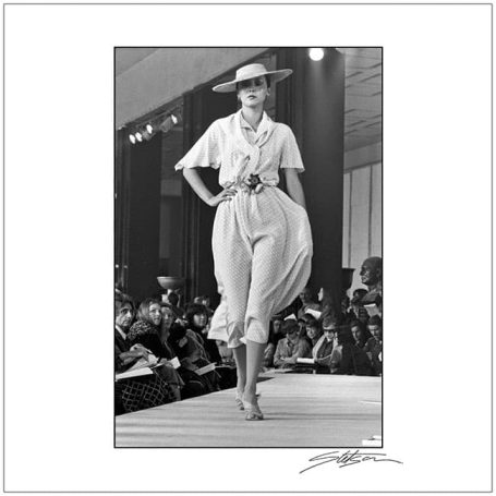 Model walking down a runway in a stylish outfit and hat, with an audience in the background.