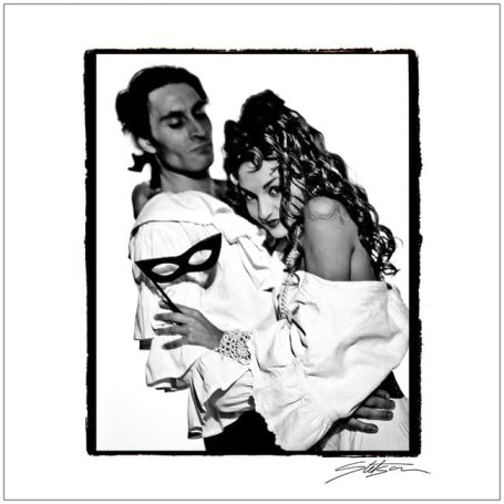 A man in a mask and a woman in a white outfit embrace, both with styled hair.