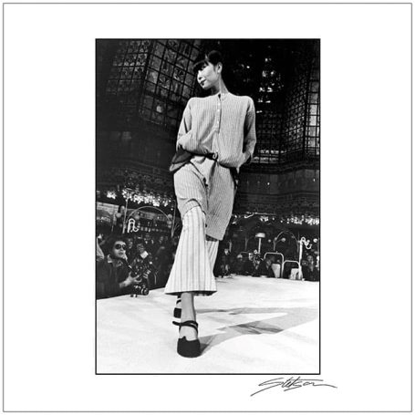 Model walking down the runway in stylish attire, set in a bustling fashion venue.