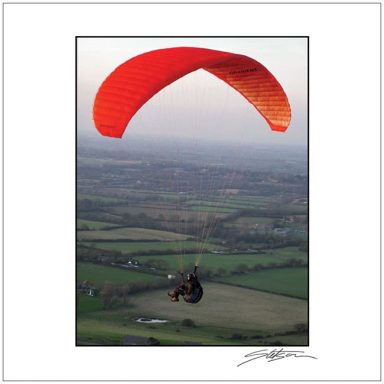 A person paragliding with a red canopy over green fields.