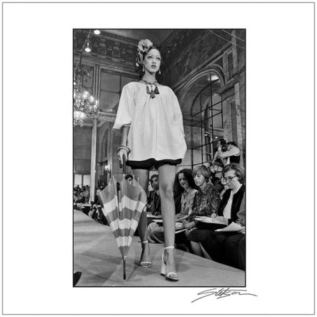 Model walking on a runway, wearing a white top and black shorts, holding an umbrella.