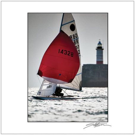 Sailing boat with a red sail near a lighthouse on a sunny day.