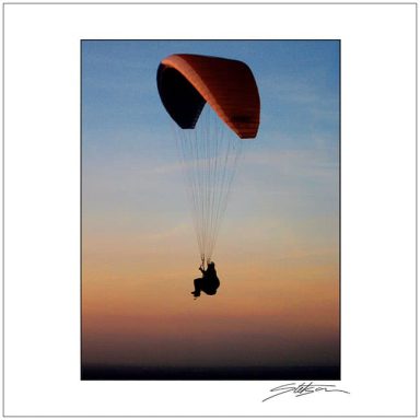 Silhouette of a person paragliding against a sunset sky with orange hues.