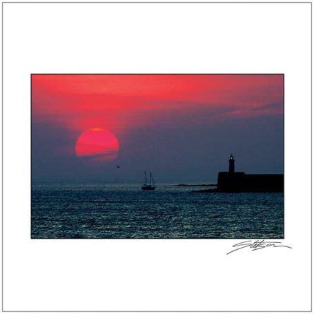 Sunset over the sea with a silhouette of a lighthouse and a sailboat in the distance.