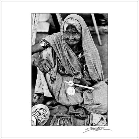 Elderly woman in traditional clothing, seated with tools and wares in a market setting.