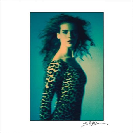 A model in a leopard print dress with flowing hair against a blue-green background.