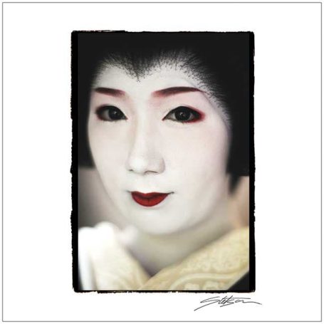 A close-up of a woman with traditional Geisha makeup, featuring white face, red lips, and dark eyes.