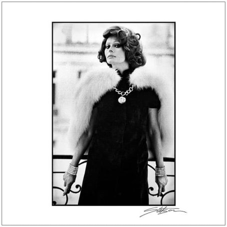 A woman in a glamorous outfit with a fur stole, standing elegantly by a balcony.