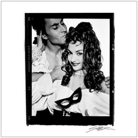 A couple in vintage attire, posing closely, with one holding a mask.
