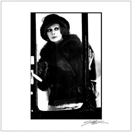 A woman in a vintage fur coat and hat stands near a door, looking outside.